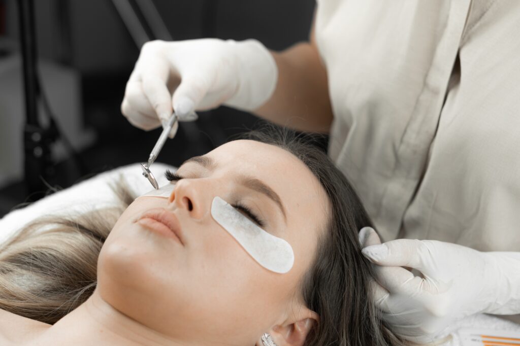 A beautician looks in a mini mirror and looks at the newly extended eyelashes of a young woman in a